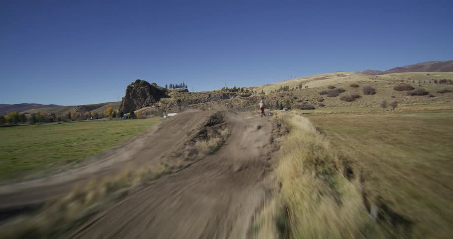 A thrilling FPV drone chases motocross riders as they speed and jump along a rural dirt track on a sunny day