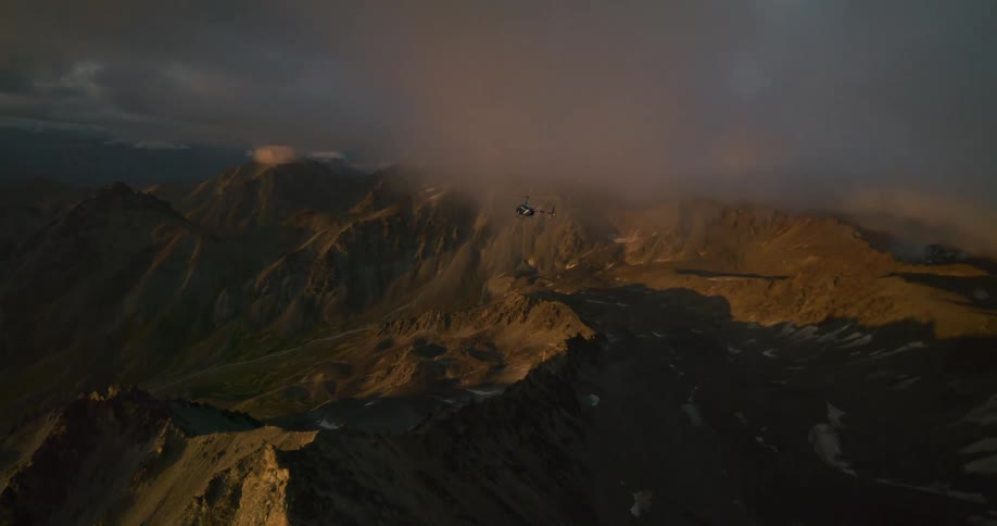 Epic aerial shot follows a helicopter flying into moody clouds over a vast mountain range at golden hour
