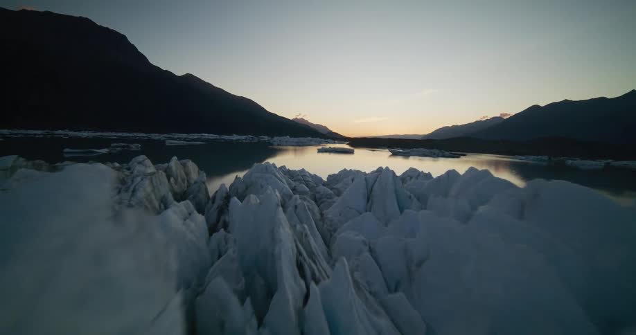 Majestic aerial footage slowly flies over the jagged, icy surface of a massive glacier towards a calm lake surrounded by mountains at dusk