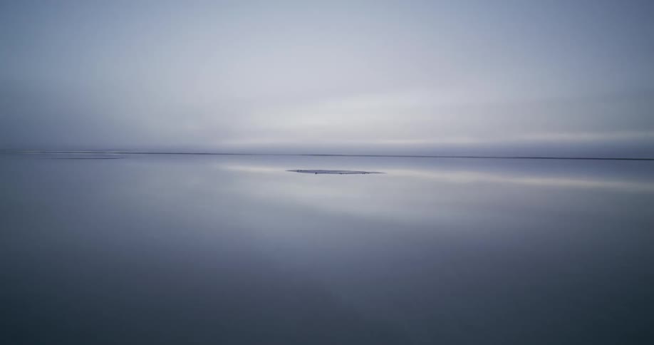 An ethereal aerial shot glides low over a vast, mirror-like body of water, where the still surface perfectly reflects the serene, overcast sky