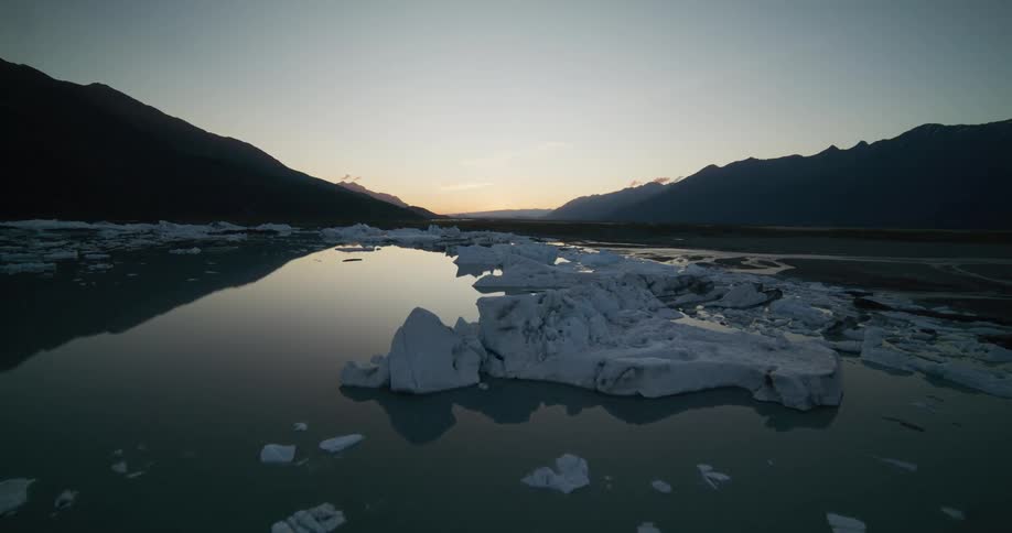 A serene aerial shot flies low over a tranquil glacial lake filled with icebergs as the last light of sunset fades behind silhouetted mountains