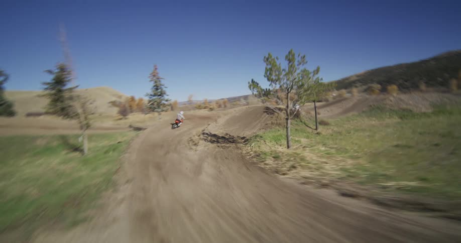 FPV drone follows motocross riders in slow motion as they race and jump on a dirt track in a sunny, rural landscape