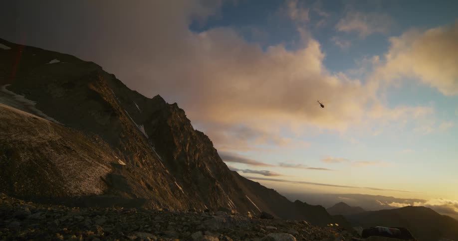 A helicopter flies away over a dramatic and rocky mountain landscape during a golden hour sunset