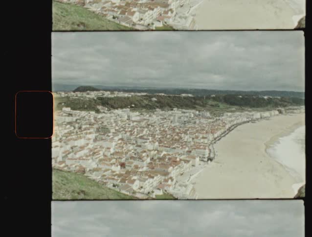 A vintage film captures the panoramic view of a coastal city with its white buildings and a long sandy beach on a cloudy day