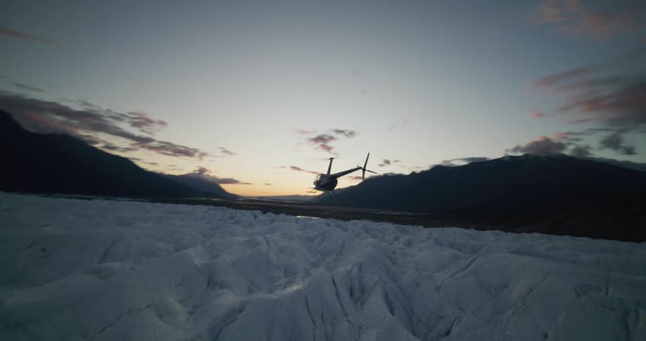 Dramatic slow-motion footage follows a helicopter flying low over a rugged glacier landscape at twilight