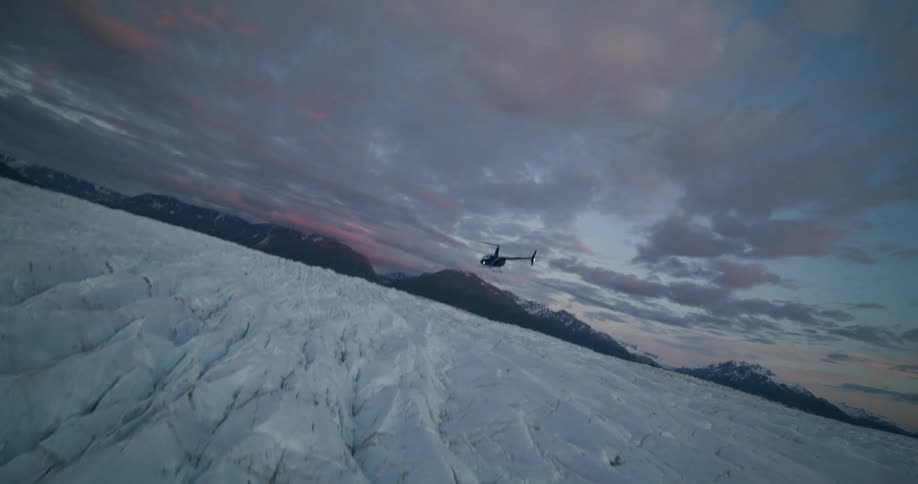 A helicopter flies low over a massive glacier field under a dramatic sunset sky in this tilted aerial shot