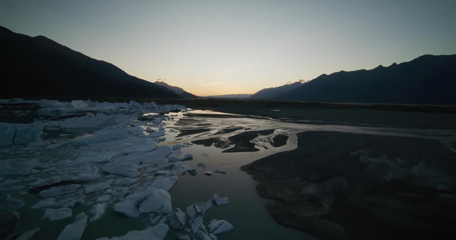 Aerial footage glides over a serene glacial river filled with melting ice chunks in a vast mountain valley at dusk