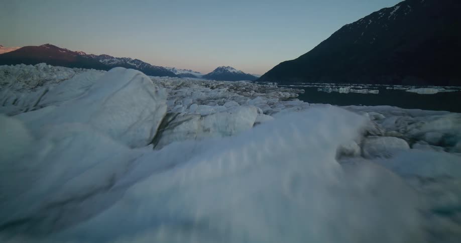 A breathtaking aerial journey over a vast, cracked glacier meeting a tranquil lake surrounded by mountains at twilight