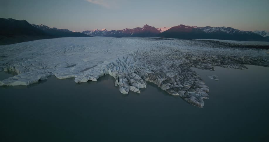 A majestic aerial flight over a calm glacial lake approaches a vast, textured glacier with a stunning mountain range illuminated by the setting sun in the background