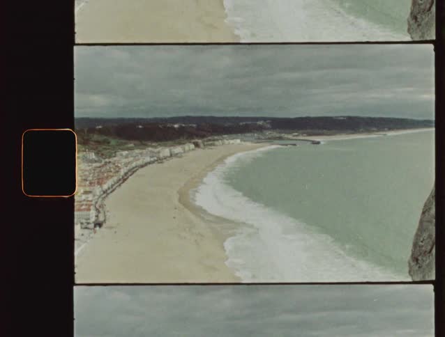 An archival film scan captures a high-angle, slow zoom-out revealing a scenic European coastal town and its long, sandy beach