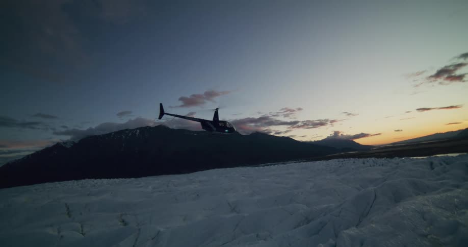 A helicopter flies low across a vast glacier and lands in the remote, mountainous wilderness at twilight