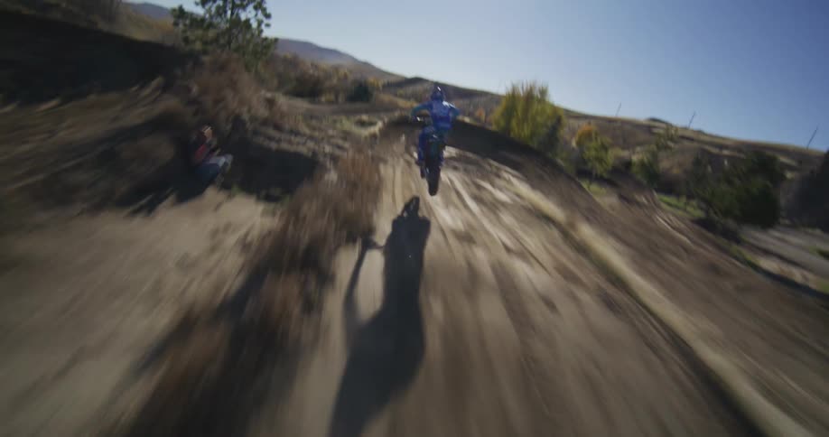 Dynamic FPV drone footage follows a motocross rider speeding through jumps and turns on a sunny dirt track