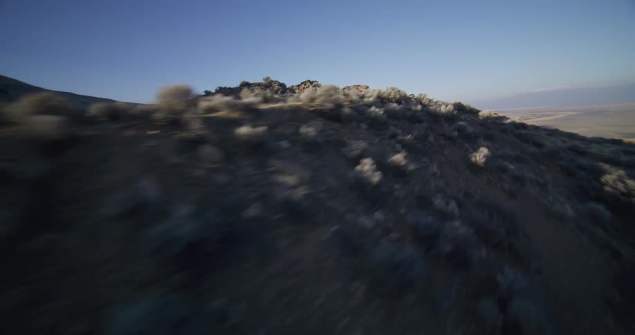 A first-person view drone flies low and fast over a rocky, sagebrush-covered mountain ridge during golden hour