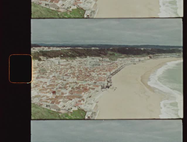A vintage 8mm film scan reveals a high-angle pan of a picturesque coastal town nestled beside a sandy beach and the ocean