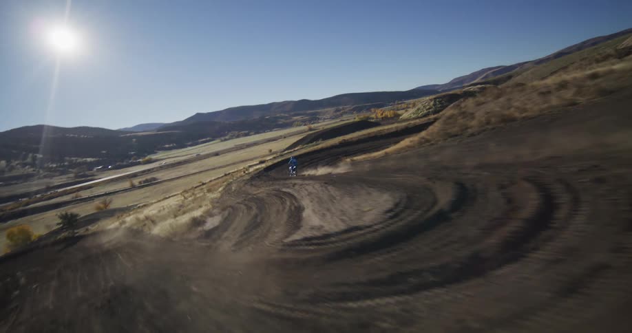 Dynamic aerial footage follows a motocross rider as they tear around a winding hillside dirt track, kicking up clouds of dust in the bright sun
