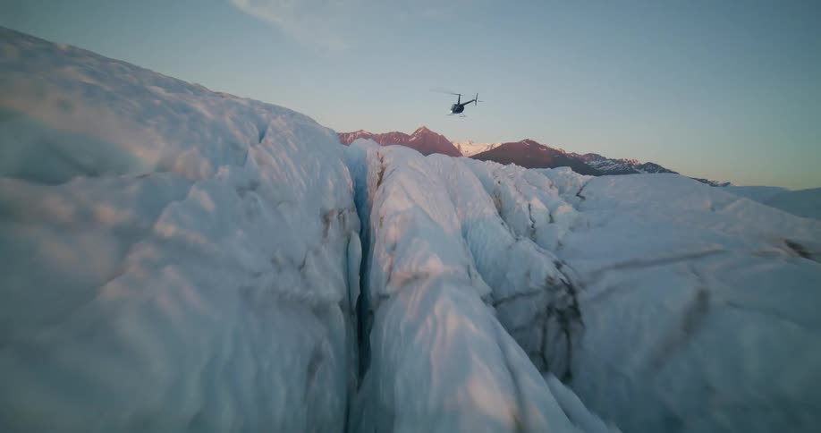 An aerial view follows a helicopter as it flies over a vast and deeply crevassed glacier towards a distant mountain range at sunset
