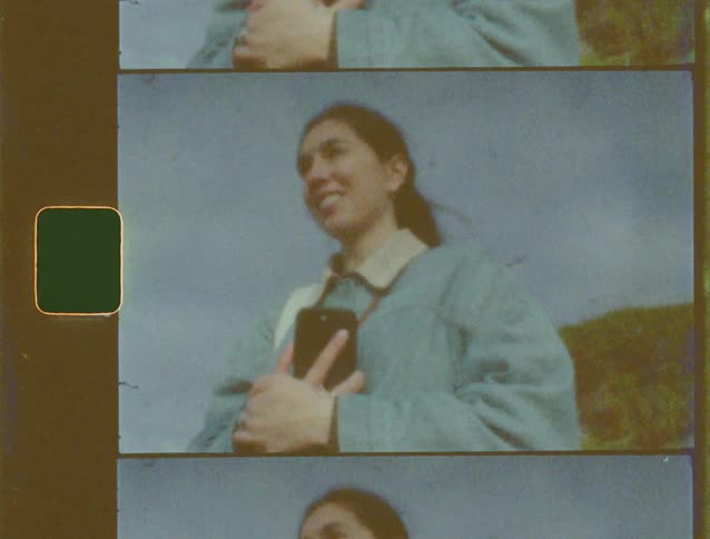 Low angle shot of a smiling young woman holding a camera outdoors on a windy day, scanned from 8mm film with vintage effects