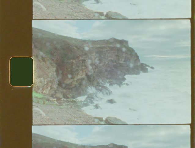 Vintage film footage shows powerful ocean waves crashing against a rugged cliffside on an overcast day