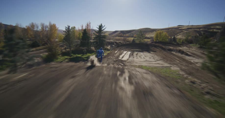 An FPV drone chases a motocross rider in slow motion as they hit a massive jump and then carve around a dusty berm on a sunny day