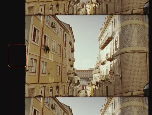 A vintage film-style shot tilts down a narrow, sunlit street in Lisbon, showing colorful tiled apartment buildings