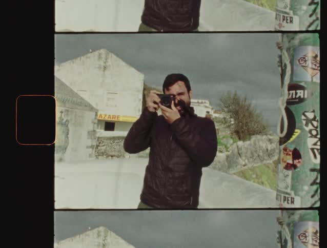 Vintage 8mm film scan of a bearded man smiling while taking a photo in a scenic European village