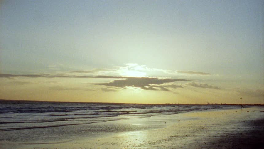 A serene, grainy film captures a hazy sunset over a wide, sandy beach as waves roll onto the shore