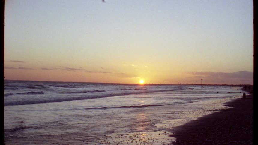 A nostalgic, grainy film captures the golden sun setting over the ocean as gentle waves lap against the shore