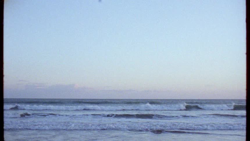 Calm ocean waves roll towards the shore under a pastel sky at dusk in a vintage film shot