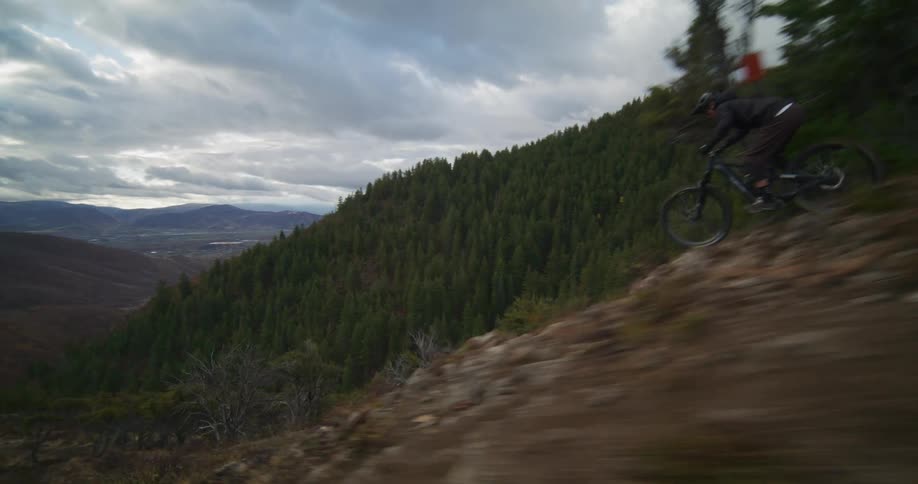 A dynamic following shot shows a mountain biker descending a steep and technical ridgeline trail in a vast mountain valley