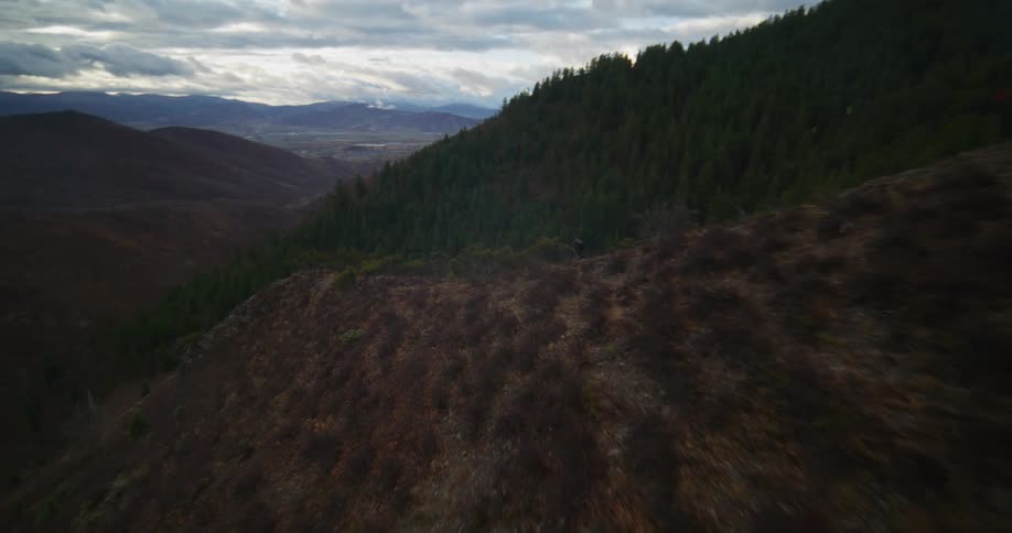 A dynamic aerial shot follows a person mountain biking along a narrow ridge trail through a vast mountain valley on a cloudy day