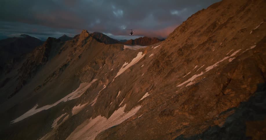 A cinematic aerial shot follows a helicopter as it soars over a rugged, snow-dusted mountain range during a dramatic sunset