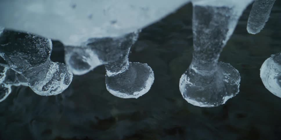 A slow-motion close-up shot reveals uniquely shaped icicles hanging just above the surface of a dark, flowing stream