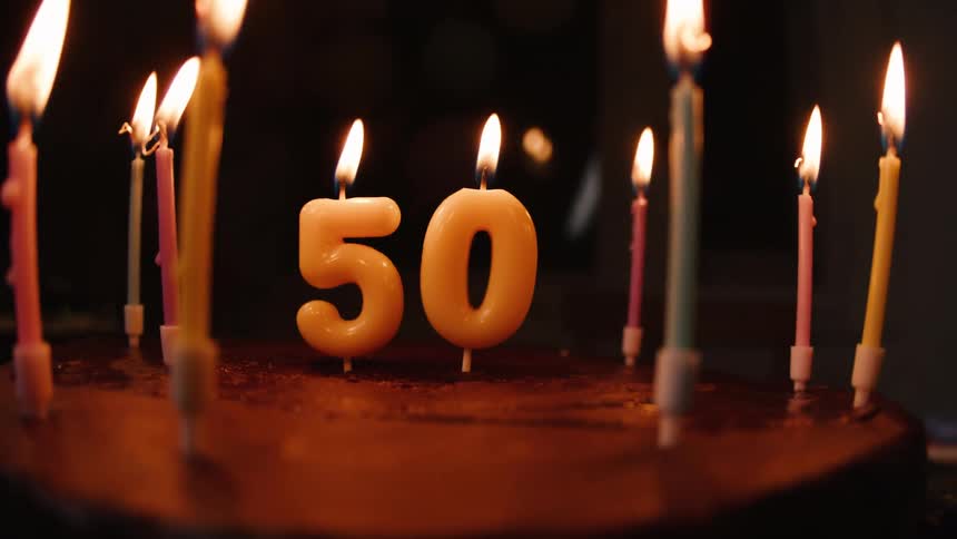 A chocolate birthday cake with the number 50 and other colorful candles lit for a celebration in a dark room