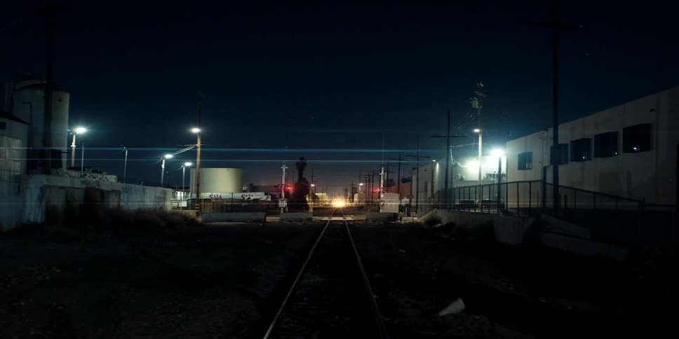 Nighttime view of a train crossing with flashing warning lights in a dark industrial zone