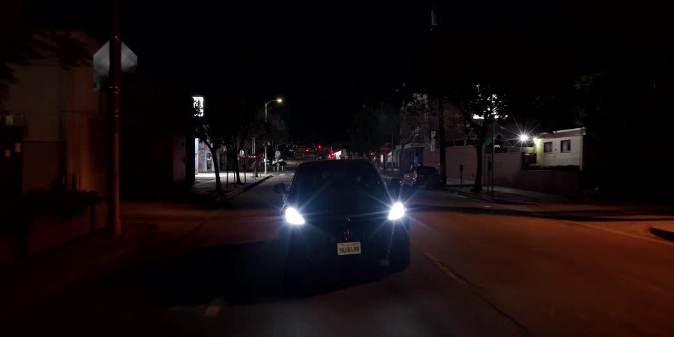 Drone view slowly pulls away from a black car parked in the middle of a deserted city street with bright headlights on at night