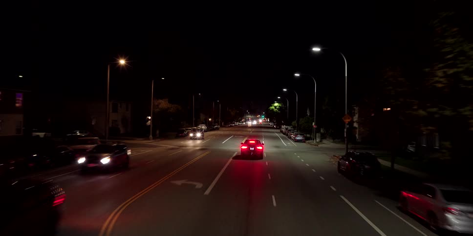 An aerial view follows a black car as it drives down a quiet, illuminated city street late at night