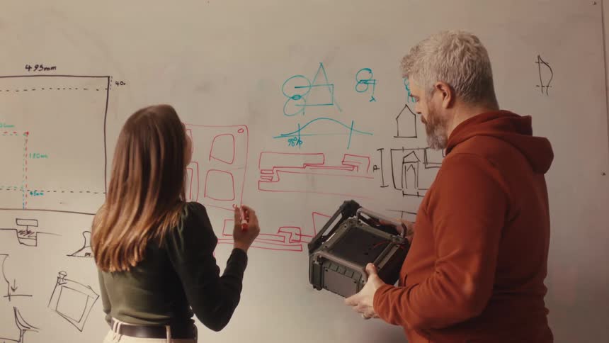 Male and female industrial designers brainstorm, comparing a physical prototype to the schematics drawn on a whiteboard
