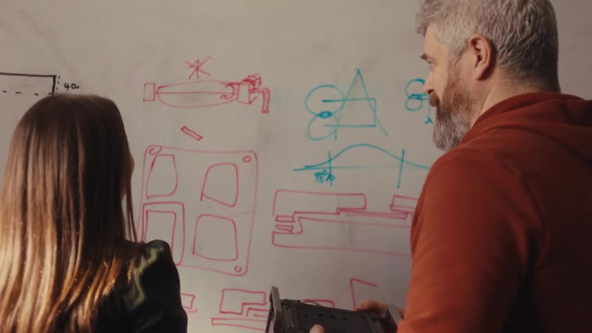 Male and female engineers discuss a product prototype while looking at technical drawings on a whiteboard