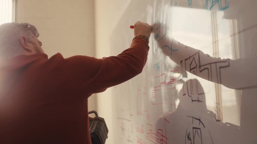 A senior engineer, backlit by a bright window, draws a colorful schematic on a whiteboard during a planning session