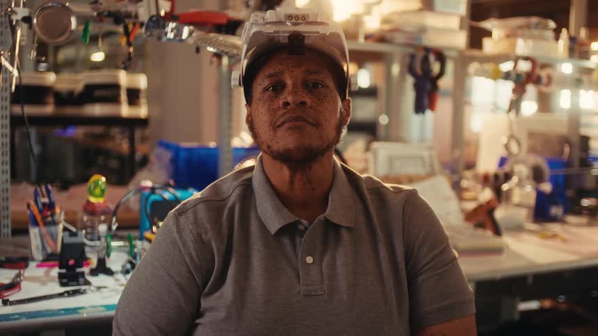 A dedicated engineer wearing a magnifying headlamp opens their eyes and stares confidently into the camera inside a cluttered electronics workshop