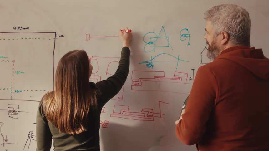 A young female engineer collaborates with her senior male colleague, drawing design schematics on a whiteboard during a brainstorming session in a creative workshop