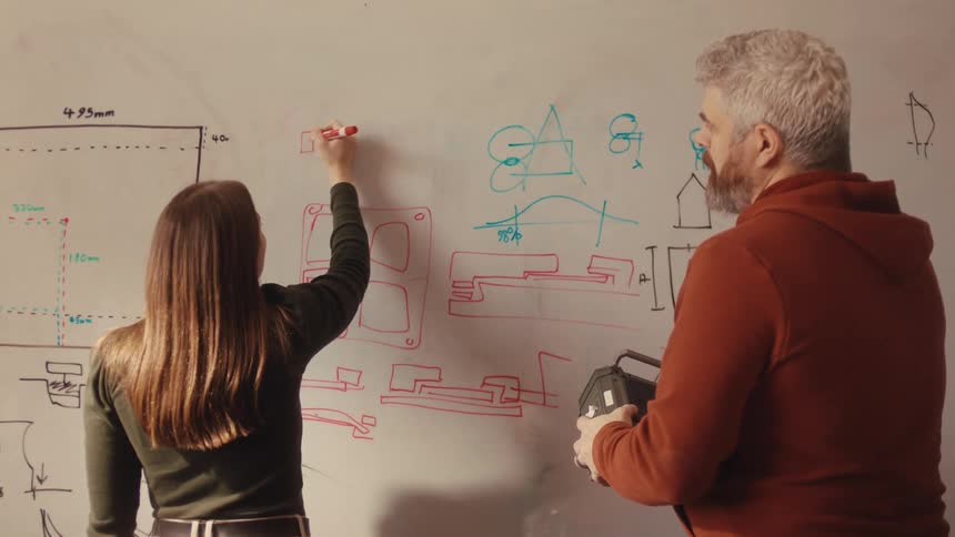 A male and female engineer collaboratively work on a technical design, drawing complex schematics on a whiteboard