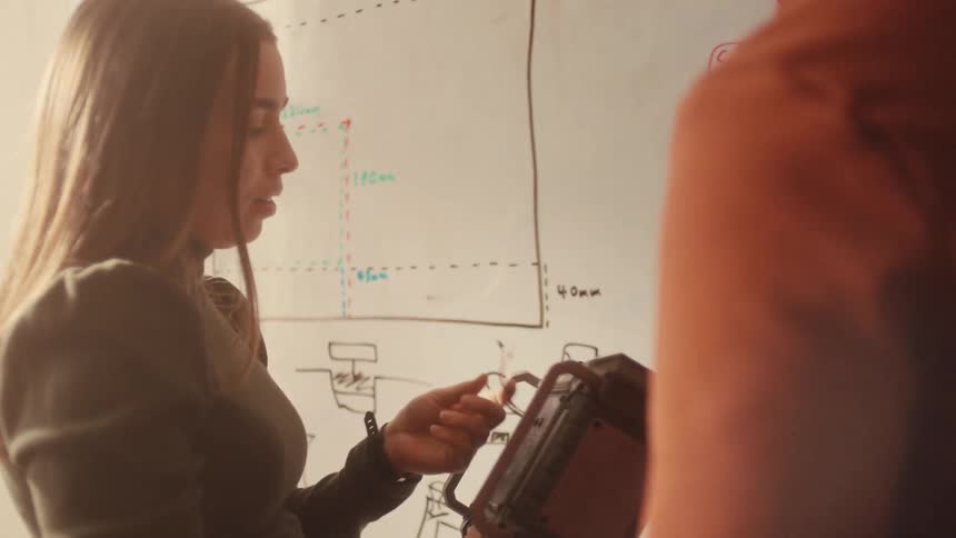 A young female engineer passionately discusses a new hardware prototype in a workshop while referencing schematics on a whiteboard