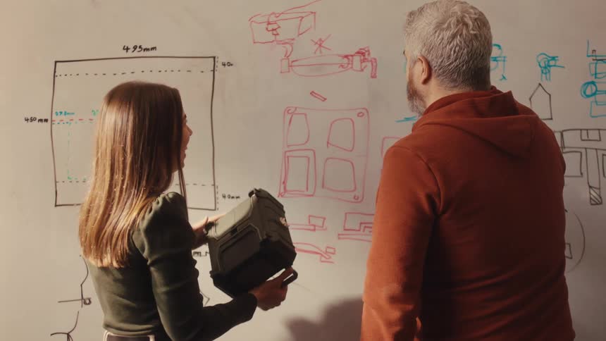 A male and female designer collaborate on a new product, sketching ideas on a whiteboard while examining a physical prototype