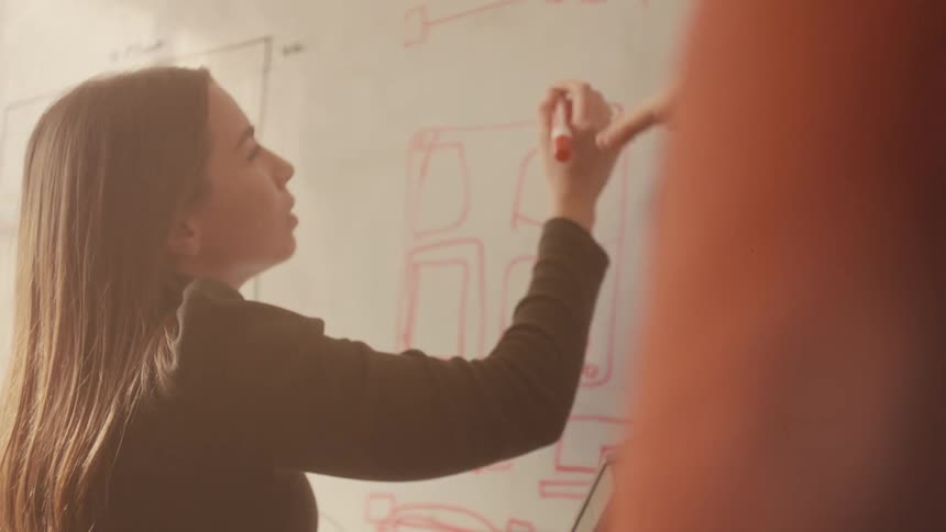 A young female engineer sketches technical designs on a whiteboard during a collaborative brainstorming session