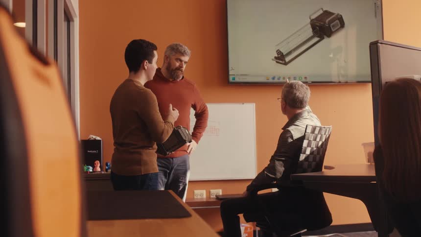 A team of male engineers collaborates in slow motion, discussing a physical prototype while viewing a 3D model on a screen in a modern office