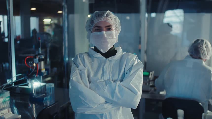 A confident female scientist in a full protective suit and mask crosses her arms while standing in a modern research laboratory