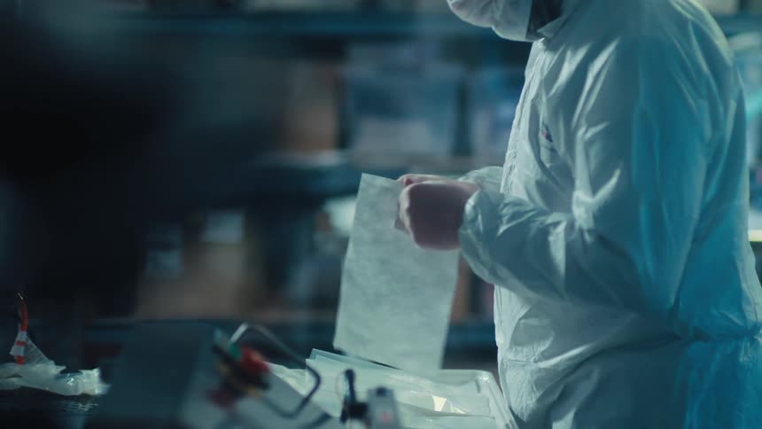 A worker in a white hazmat suit carefully unpacks a sterile sheet in a cleanroom laboratory setting