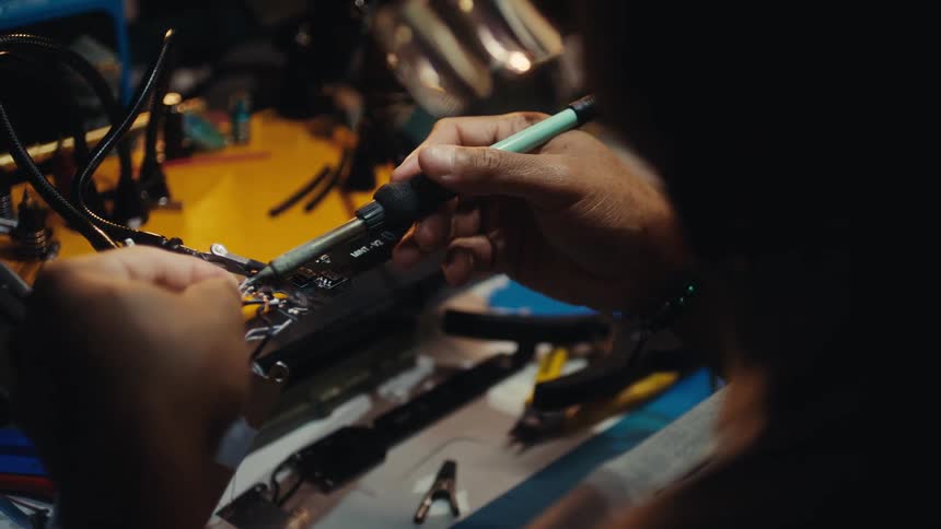 Over the shoulder shot of a technician carefully using a soldering iron to connect wires to an electronic circuit board