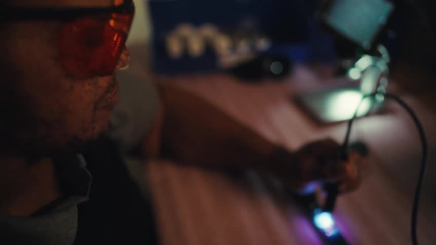 A technician in safety glasses carefully inspects an electronic component with a UV light in a dimly lit workshop